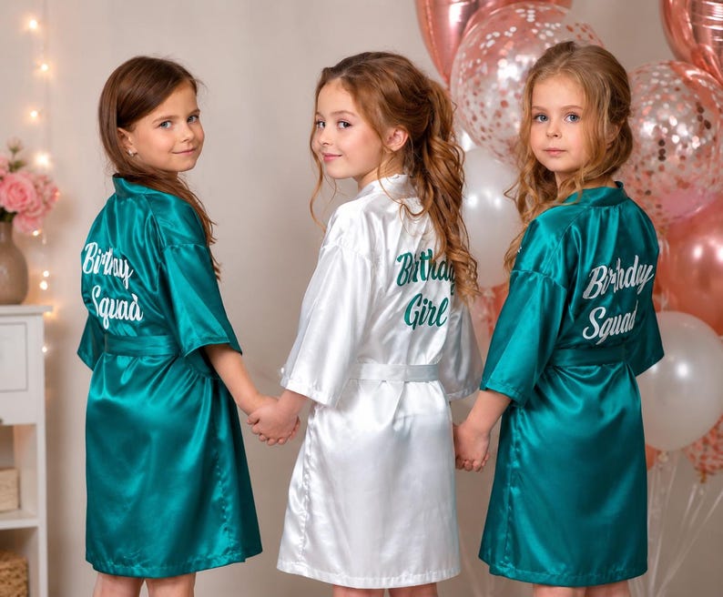 May include: Three young people wearing satin robes. Two robes are teal and say "Birthday Squad" in white script on the back. The third robe is white and says "Birthday Girl" in green script. Balloons are in the background.