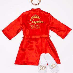 A red satin robe with gold text and a gold crown, along with white slippers with gold text that says "Mrs. Sophia".