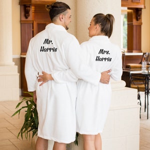 May include: Two white bathrobes with black embroidery. The robe on the left says "Mr. Harris" and the robe on the right says "Mrs. Harris".