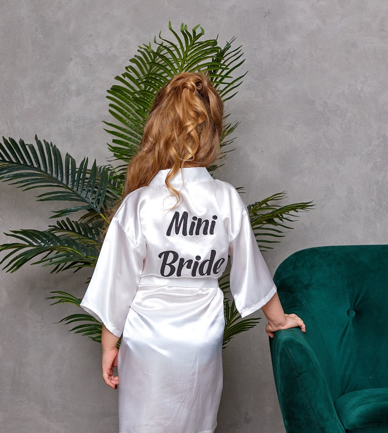 May include: A white satin robe with the words "Mini Bride" printed in black on the back. The robe is worn by a person with long, wavy blonde hair. The background includes a green plant and a dark green armchair.