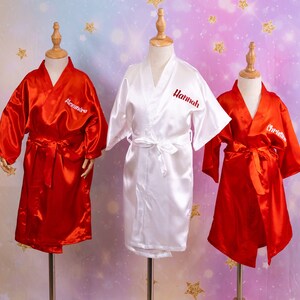 May include: Three satin robes on display. Two are red, and one is white. Each robe has a name embroidered on the front: "Alexandra", "Hannah", and "Christina". The robes have a matching belt.
