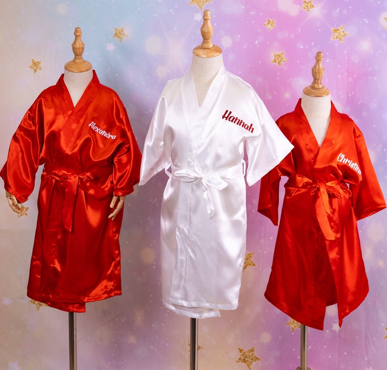 May include: Three satin robes on display. Two are red, and one is white. Each robe has a name embroidered on the front: "Alexandra", "Hannah", and "Christina". The robes have a matching belt.
