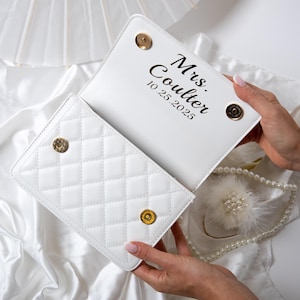 May include: White quilted clutch with gold-tone hardware. The interior features the inscription "Mrs. Coulter 10.25.2025". The clutch is open, revealing the interior and a pearl and feather accessory.