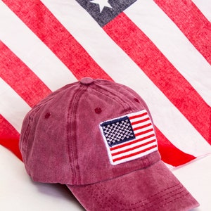 May include: A burgundy baseball cap with a small American flag patch on the front. The cap is made of a soft, distressed cotton fabric.