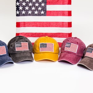 May include: Five baseball caps in different colors with an American flag patch on the front. The caps are arranged in a row on a white surface. The caps are blue, gray, yellow, red, and brown.