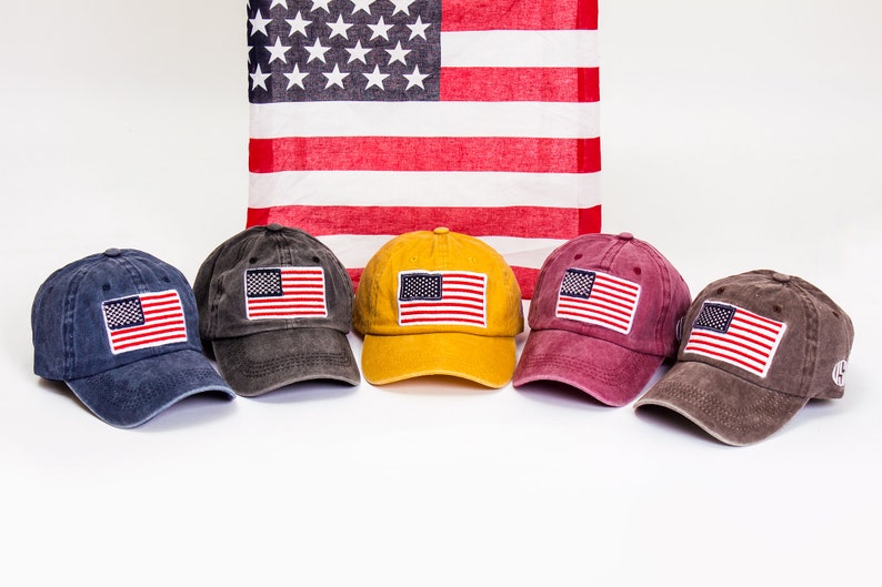 May include: Five baseball caps in different colors with an American flag patch on the front. The caps are arranged in a row on a white surface. The caps are blue, gray, yellow, red, and brown.