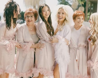 May include: A group of women in pale pink satin robes with feather trim, posing outdoors. The bride is in a white dress and veil, surrounded by bridesmaids. The robes have a soft, elegant appearance, suitable for a bridal party.