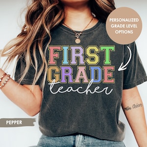 May include: A charcoal gray short-sleeve t-shirt featuring the text "FIRST GRADE" in colorful block letters and the word "teacher" in white cursive script. The shirt includes a circular graphic in the top right corner with the text "PERSONALIZED GRADE LEVEL OPTIONS" and a label in the bottom left corner reading "PEPPER".