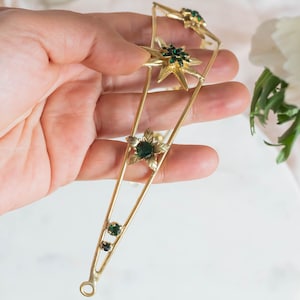 May include: A gold tiara with a pointed design features five green star-shaped gemstones. The tiara is held in a hand.