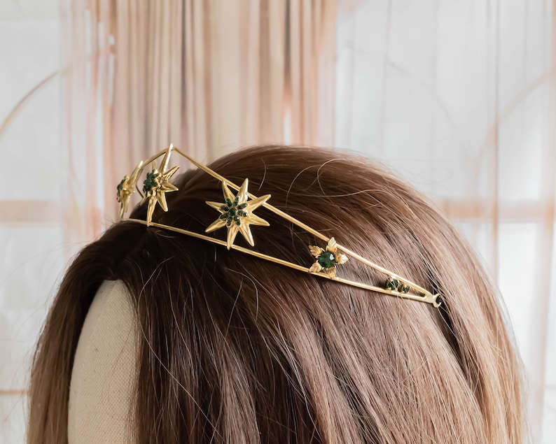 May include: A gold crown with five pointed stars and green gemstones. The crown is on a brown wig.