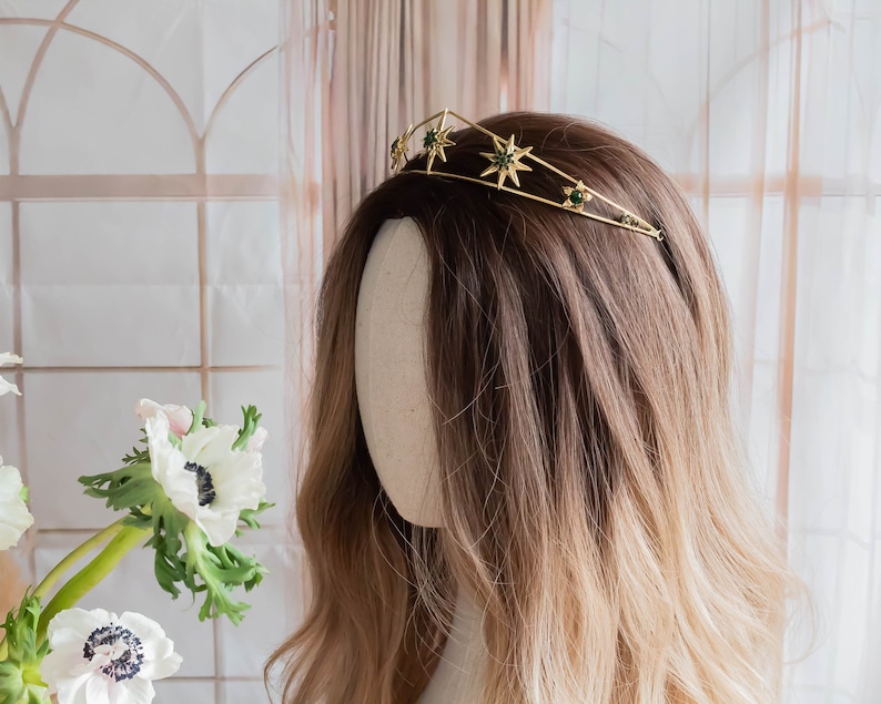May include: A gold crown with green star-shaped accents. The crown is on a mannequin head with long, brown and blonde hair.