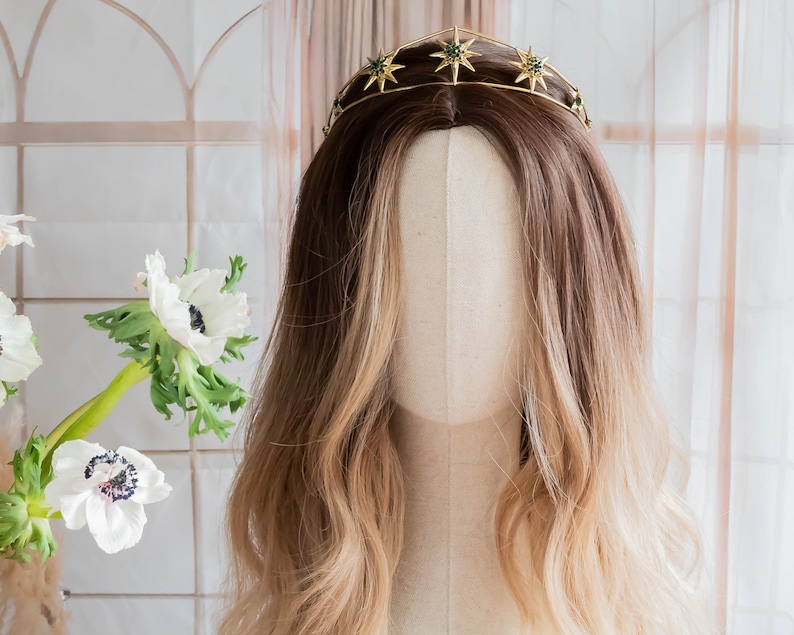 May include: A gold crown with green star accents. The crown is on a mannequin head with long, wavy, light brown hair.