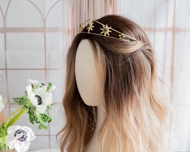May include: Gold star crown with green gemstones, worn on a mannequin with long brown and blonde hair.