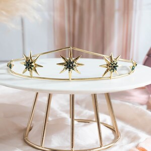 May include: A gold crown with five pointed star details, each star has a dark green center. The crown is sitting on a white cake stand with a gold metal base.