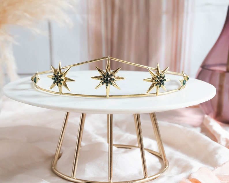 May include: A gold crown with five pointed star details, each star has a dark green center. The crown is sitting on a white cake stand with a gold metal base.