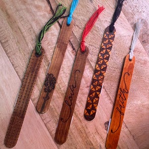May include: Five wooden bookmarks with different designs. One bookmark has a flower design, another says "love", another has a geometric pattern, another says "faith", and the last one has a dotted pattern.
