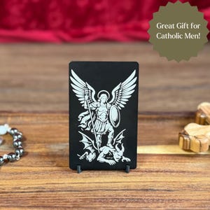 May include: Black rectangular plaque with a white illustration of Saint Michael the Archangel, holding a spear and shield, standing over a demon. The text "Great Gift for Catholic Men!" is in a green starburst shape.