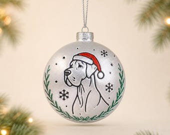May include: A silver spherical Christmas ornament featuring a black line drawing of a Great Dane dog wearing a red Santa hat. The design is framed by green leafy branches and small black snowflakes, suspended by a silver string loop.