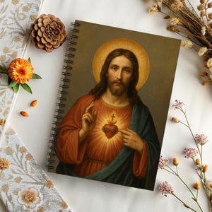 Puede incluir: Un cuaderno de espiral que presenta una ilustración religiosa clásica de Jesucristo con un halo dorado, vistiendo una túnica roja y un manto azul, con un Sagrado Corazón resplandeciente en el centro del pecho y una mano en bendición.