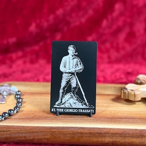 May include: A black and white image of St. Pier Giorgio Frassati, standing with a walking stick and a pipe in his mouth. The image is on a black card with the text "ST. PIER GIORGIO FRASSATI" at the bottom. The card is displayed on a wooden surface.