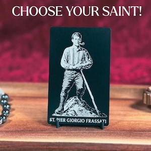 May include: A black rectangular plaque with a white illustration of a man holding a pipe and a staff, standing on a rock. The text "CHOOSE YOUR SAINT!" is at the top, and "ST. PIER GIORGIO FRASSATI" is at the bottom.