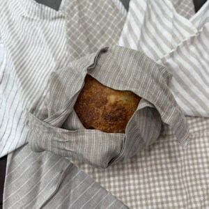 May include: A loaf of freshly baked bread wrapped in a beige and white striped linen cloth. Several other linen cloths with various patterns, including stripes and checks, are visible in the background. The bread has a golden-brown crust.