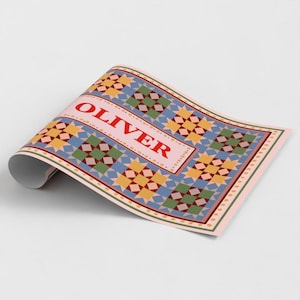 May include: A rectangular sheet of paper featuring a repeating quilt-like pattern of yellow, green, and blue squares. The center contains a light pink rectangular field with the name OLIVER printed in bold red capital letters.