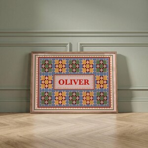 May include: A rectangular framed print featuring a repeating quilt-style pattern of blue, yellow, and green star motifs. The center contains a light pink rectangular panel with the name OLIVER printed in bold red capital letters.