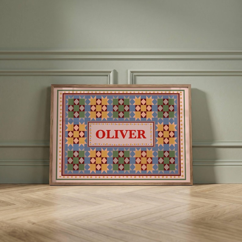 May include: A rectangular framed print featuring a repeating quilt-style pattern of blue, yellow, and green star motifs. The center contains a light pink rectangular panel with the name OLIVER printed in bold red capital letters.