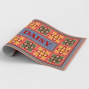 May include: A roll of wrapping paper featuring a repeating quilt-like pattern of yellow, green, and red geometric shapes on a coral background. A central blue horizontal band displays the word DAISY in bold red capital letters.