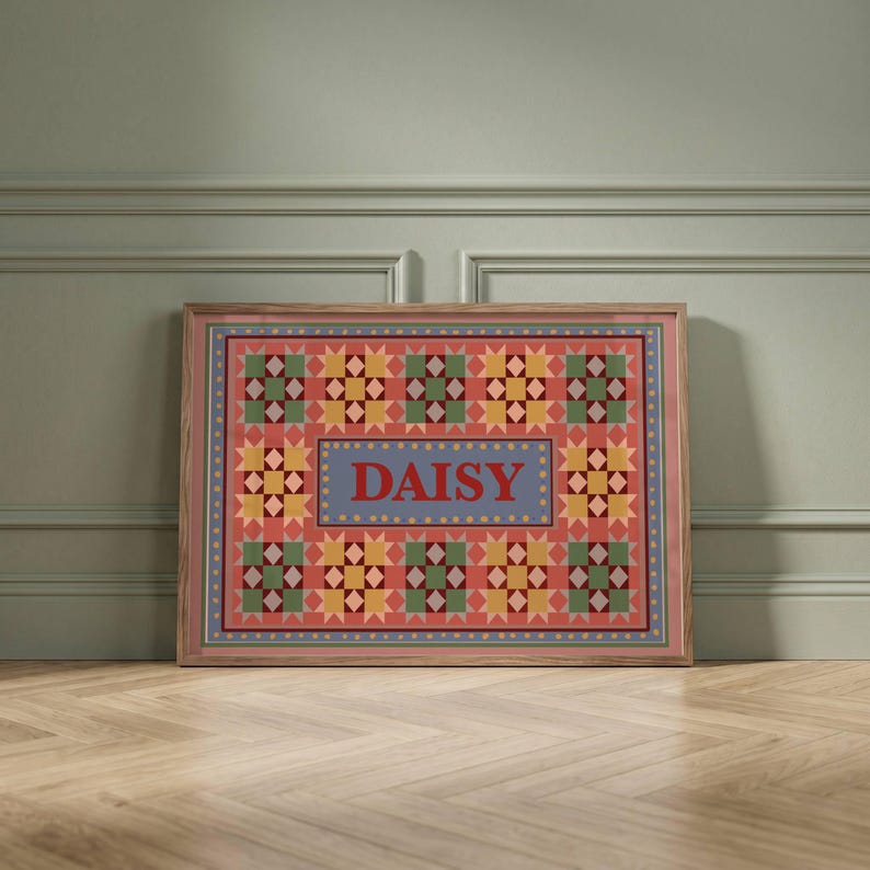 May include: A rectangular framed print featuring a quilt-inspired pattern with a repeating star motif in shades of red, yellow, and green. The center contains a blue rectangular box with the name DAISY printed in bold red capital letters.