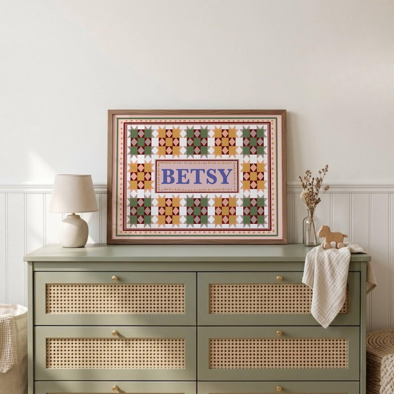 May include: A framed rectangular wall art piece featuring a quilt-inspired pattern of green, yellow, and white geometric star shapes surrounding a central rectangular panel with the name BETSY printed in large blue capital letters.
