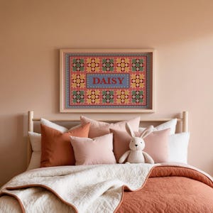 May include: A rectangular framed wall art piece featuring a quilt-inspired pattern of geometric stars in shades of pink, green, and yellow, with the name DAISY printed in red letters inside a central blue rectangle with a dotted border.