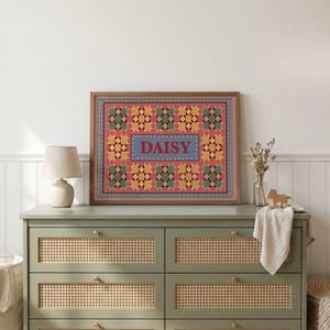 May include: A rectangular framed wall art piece featuring a repeating geometric star pattern in shades of pink, yellow, green, and blue. The center contains a blue rectangular panel with the name DAISY printed in bold red capital letters.