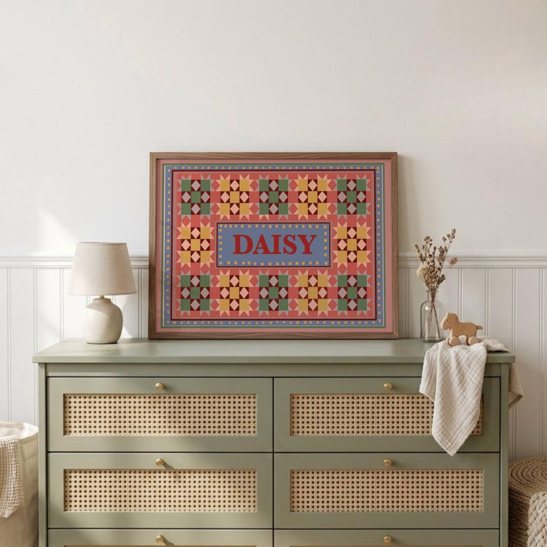 May include: A rectangular framed wall art piece featuring a repeating geometric star pattern in shades of pink, yellow, green, and blue. The center contains a blue rectangular panel with the name DAISY printed in bold red capital letters.