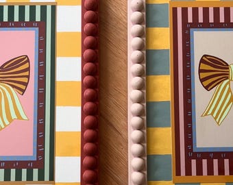 May include: Two decorative picture frames. One frame is burgundy with a row of round accents, and the other is light pink with similar accents. Both frames have a yellow and white striped background and a pink center with a bow illustration.
