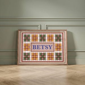 May include: A rectangular framed print featuring a repeating geometric quilt pattern in shades of green, yellow, and maroon. The center contains a light pink rectangular field with the name BETSY printed in large, bold, blue serif letters.