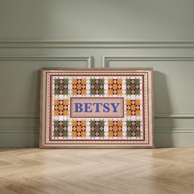 May include: A rectangular framed print featuring a repeating geometric quilt pattern in shades of green, yellow, and maroon. The center contains a light pink rectangular field with the name BETSY printed in large, bold, blue serif letters.