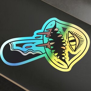 May include: A rainbow holographic sticker of a cartoon mushroom with an eye and dripping liquid. The sticker is on a black skateboard.