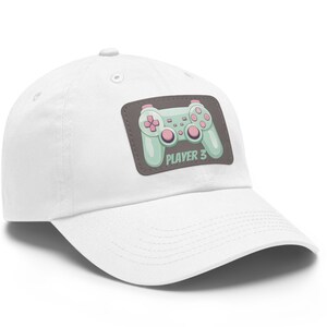 Op de afbeelding: Witte baseballpet met een groene en roze gamepad-afbeelding en de tekst "PLAYER 3" op een bruine patch.