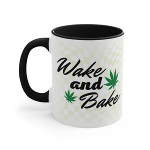 May include: White ceramic mug with a black rim and handle. The mug has a checkered pattern and the text "Wake and Bake" with green marijuana leaf illustrations.