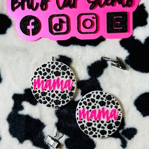 May include: Two white car air fresheners with a black and white animal print design. The air fresheners have the word "mama" written in pink glitter.