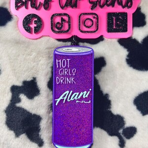 Op de afbeelding: Paarse en blauwe autoluchtverfrisser in de vorm van een blikje Alani Nu energiedrank. Het blikje heeft de tekst "Hot Girls Drink Alani Nu" erop.