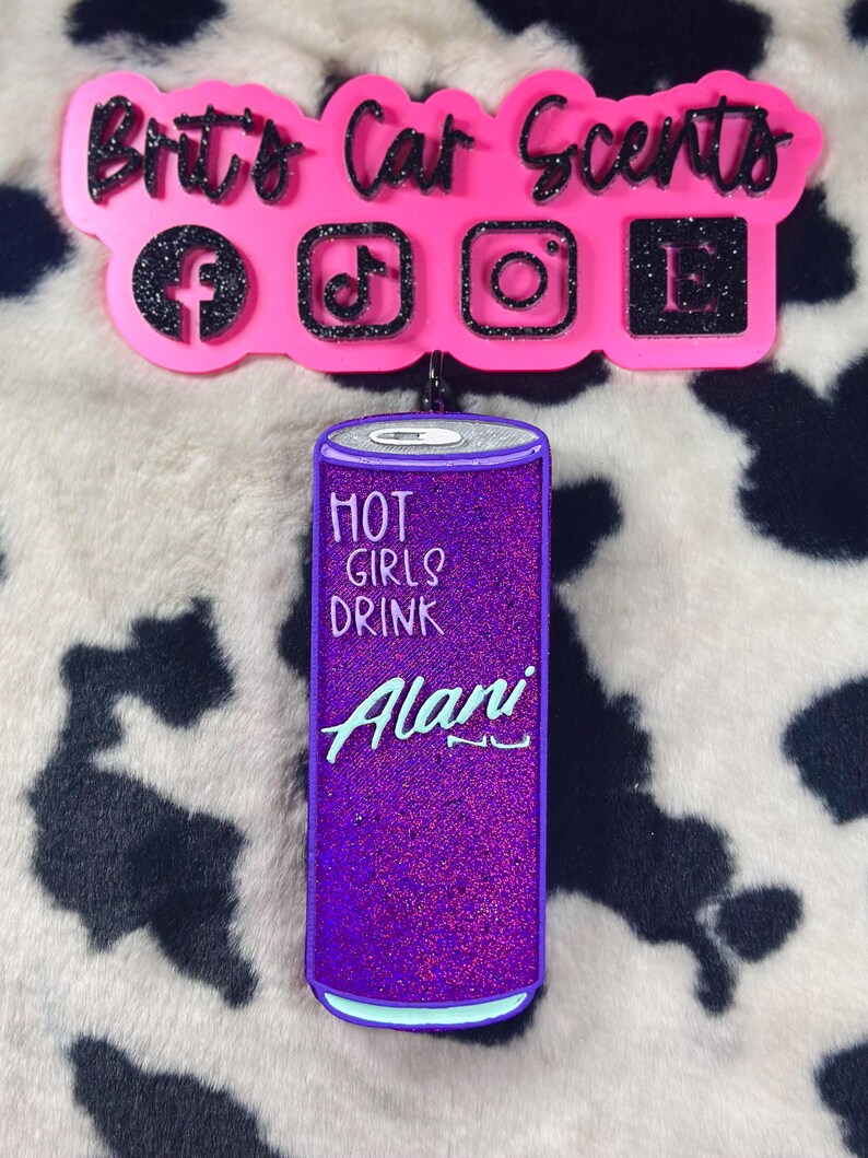 Op de afbeelding: Paarse en blauwe autoluchtverfrisser in de vorm van een blikje Alani Nu energiedrank. Het blikje heeft de tekst "Hot Girls Drink Alani Nu" erop.