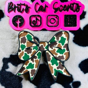 May include: Pink plastic mold with the text "Britt's Car Scents" and social media icons for Facebook, TikTok, Instagram, and YouTube. A brown, green, and white camouflage bow is in the foreground.