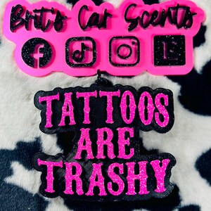 May include: Pink and black car air freshener with the text "Britt's Car Scents" and social media icons. The bottom part of the air freshener has the text "Tattoos Are Trashy" in black with pink glitter.