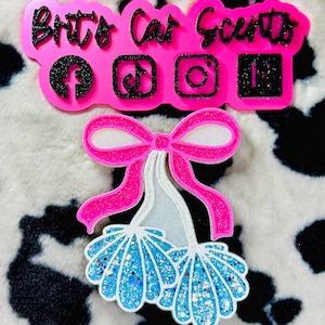 May include: A pink car scent charm with the text "Brit's Car Scents" and social media icons. Below is a bow-shaped charm with a pink bow and blue, glittery seashell-like accents. The background is a black and white cow print.