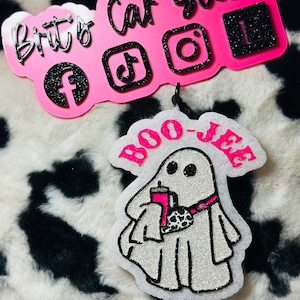 K&ouml;nnte beinhalten: Ein wei&szlig;er, glitzernder, geisterf&ouml;rmiger Auto-Lufterfrischer mit dem Text BOO-JEE in leuchtendem Pink. Der Geist h&auml;lt einen pinken Becher und tr&auml;gt eine Bauchtasche mit Kuhmuster. Dar&uuml;ber ein pinkes Schild mit schwarzem Glitzertext Brits Car Scents.