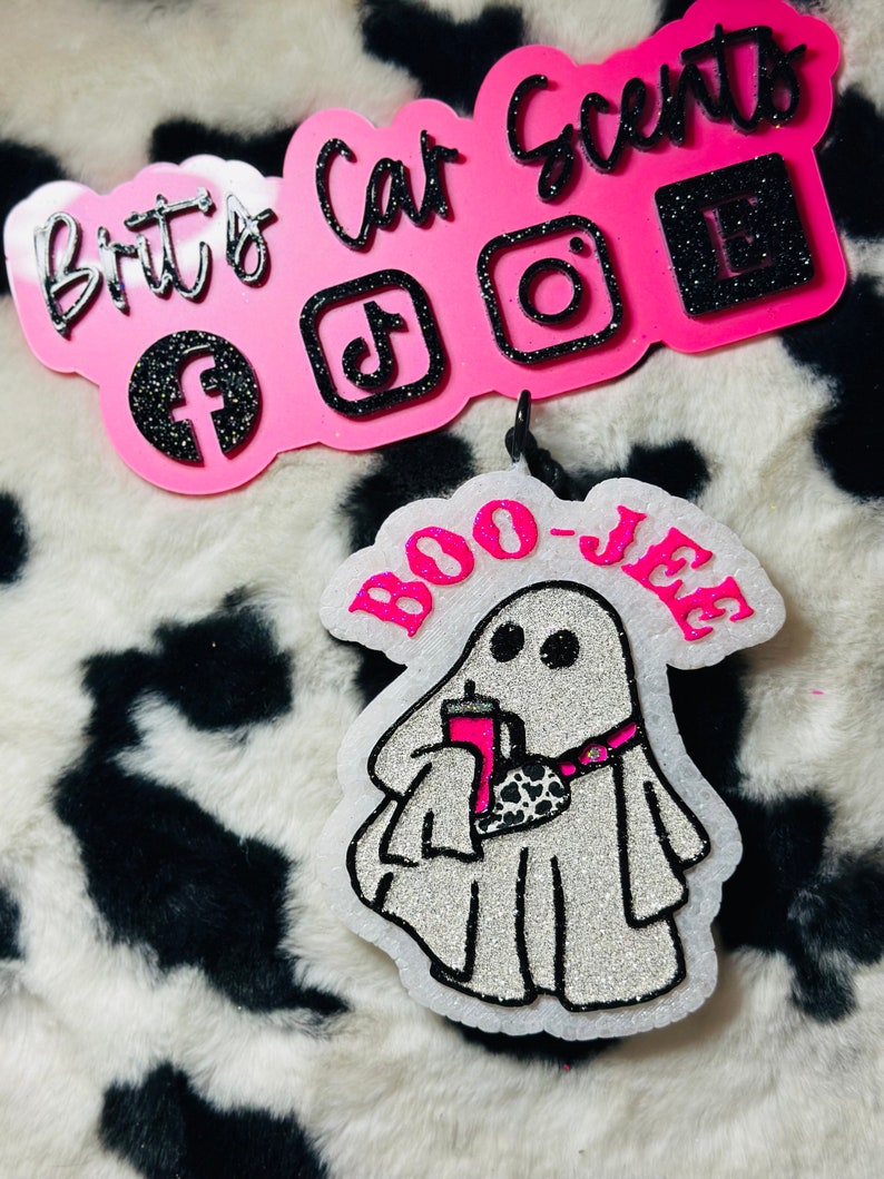 K&ouml;nnte beinhalten: Ein wei&szlig;er, glitzernder, geisterf&ouml;rmiger Auto-Lufterfrischer mit dem Text BOO-JEE in leuchtendem Pink. Der Geist h&auml;lt einen pinken Becher und tr&auml;gt eine Bauchtasche mit Kuhmuster. Dar&uuml;ber ein pinkes Schild mit schwarzem Glitzertext Brits Car Scents.