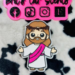 May include: A cartoon-style Jesus figure with outstretched arms, wearing a white robe and a pink sash. Above, a pink sign with the text "Brit's Car Scents" and social media icons.